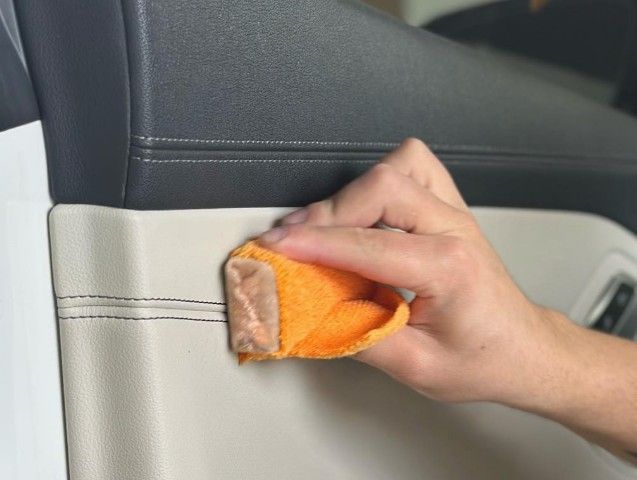 Hand cleaning car's cream-colored door panel with an orange microfiber cloth, showcasing detail.