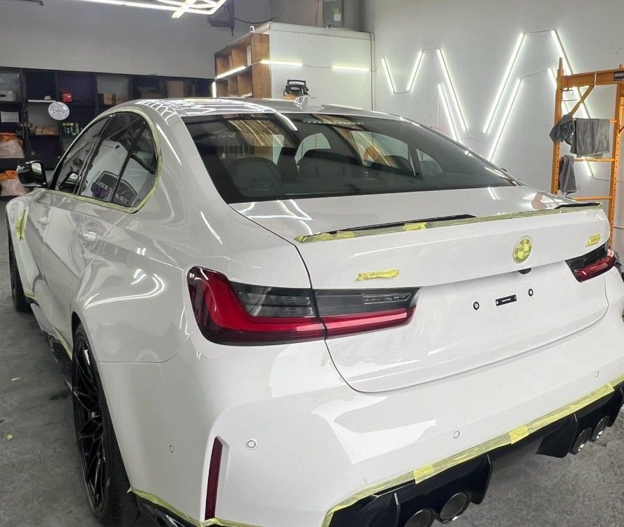 White BMW sedan being worked on in a shop. Tape outlines body panels. Black wheels.