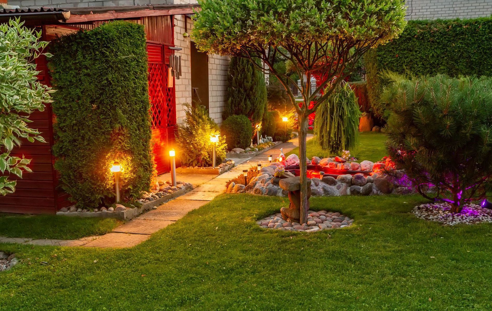 A garden at twilight features a stone path lined with warm lanterns, a manicured lawn, and a small, illuminated pond.
