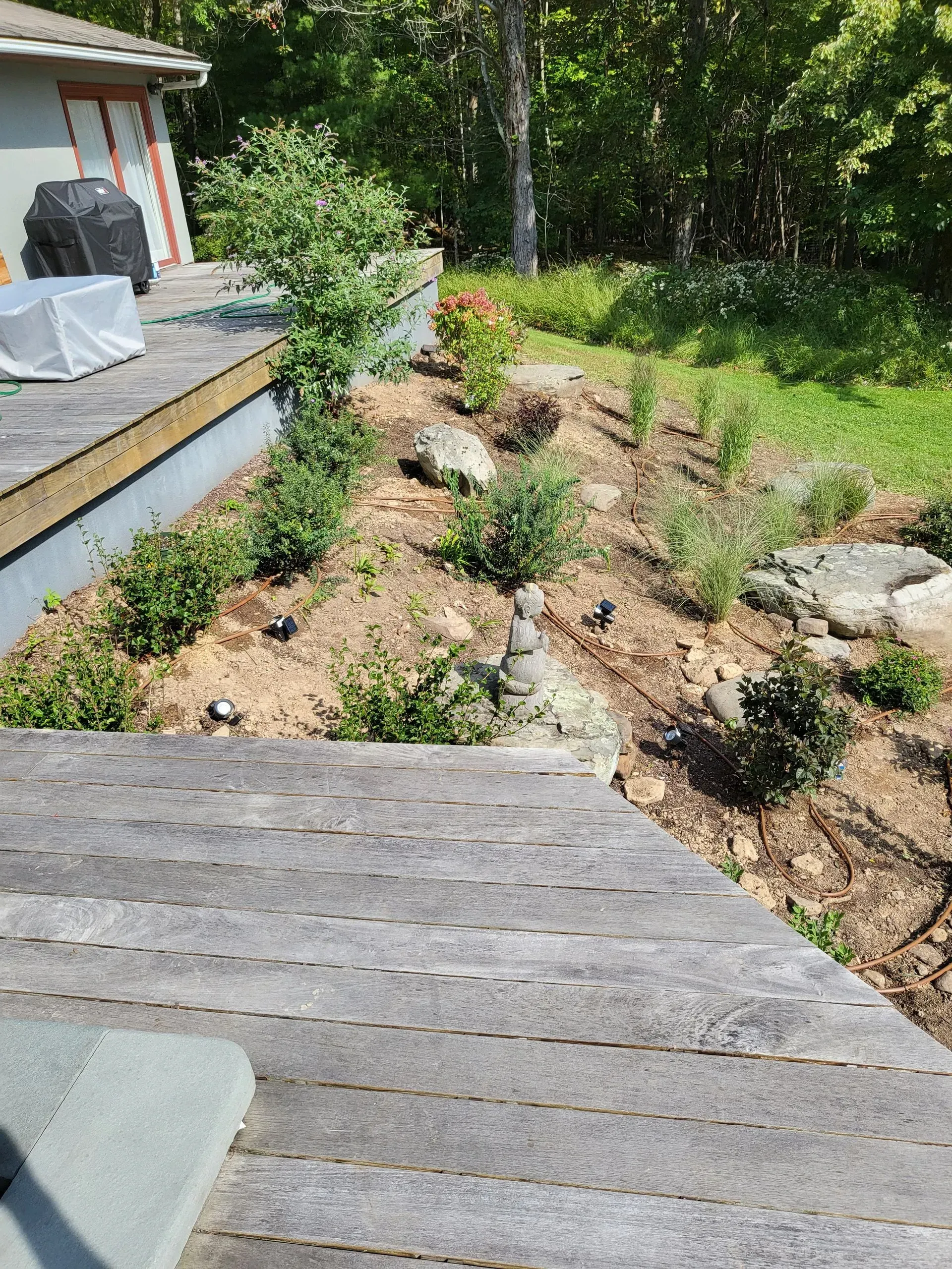 A tiered garden bed with small shrubs, stones, and a rock stack, situated next to a wooden deck.