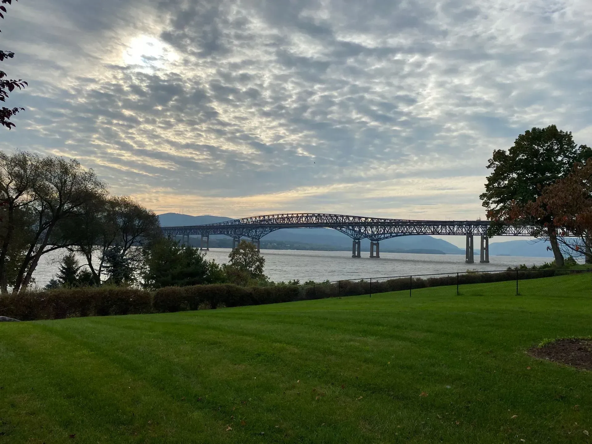A metal truss bridge spans a wide river under a cloudy sky, viewed from a grassy park area with trees on either side.