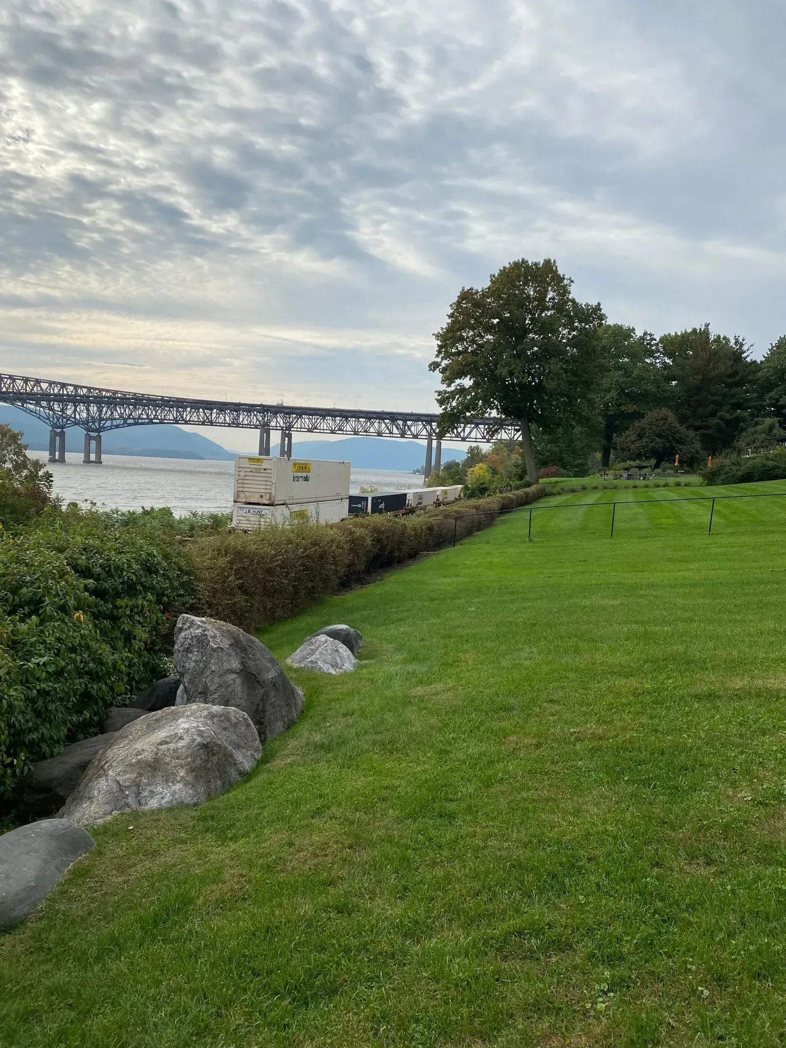 A large metal bridge spans a river behind a grassy field with large rocks and bushes in the foreground.