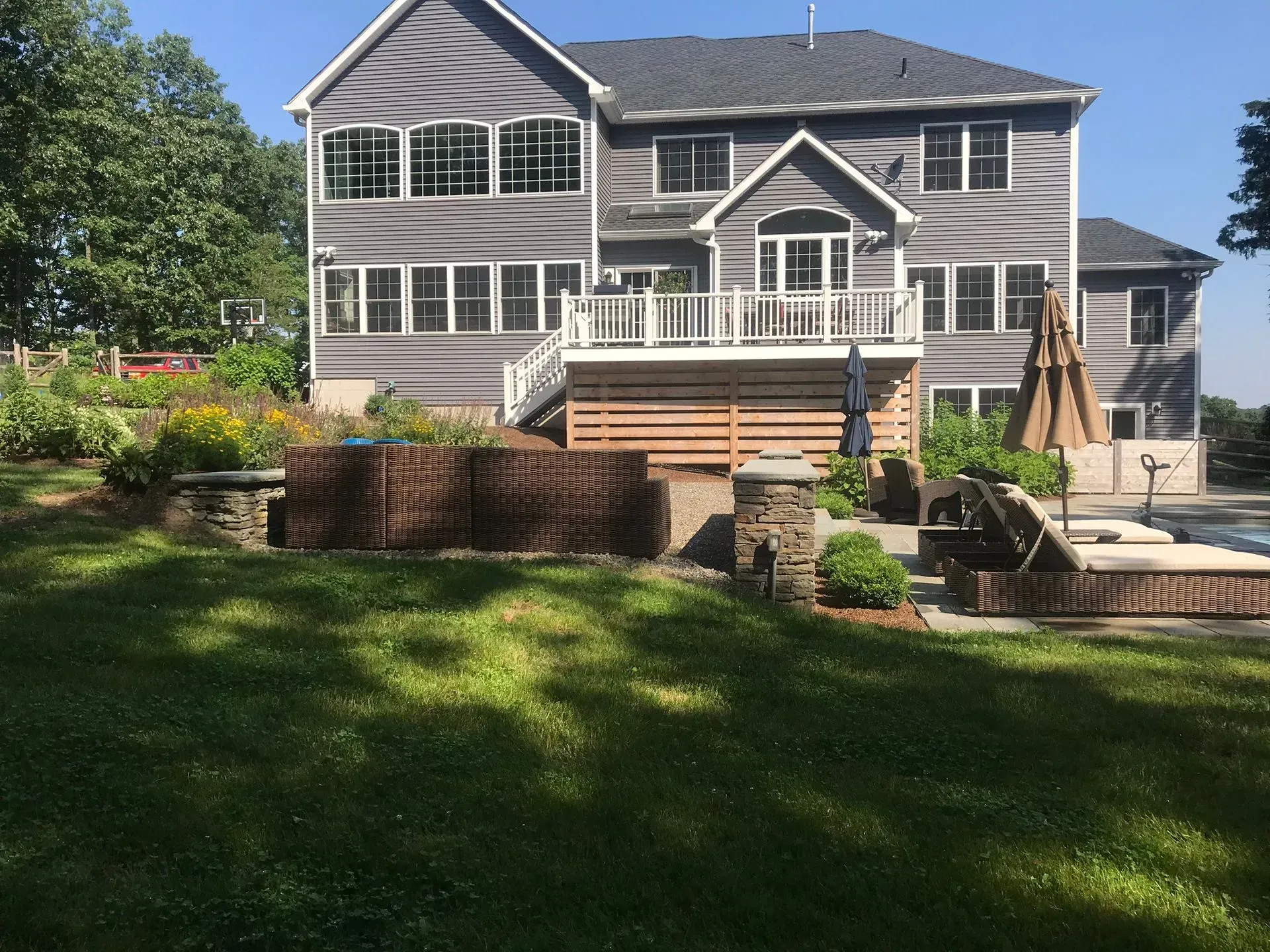 A large gray two-story house with a wooden deck, stone pillars, and a patio area overlooking a green backyard.