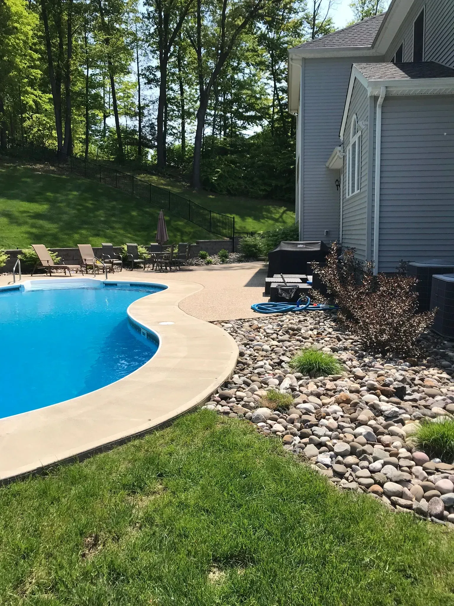A swimming pool with a curved concrete edge, surrounded by a stone patio, landscaping rocks, and a grey house exterior.