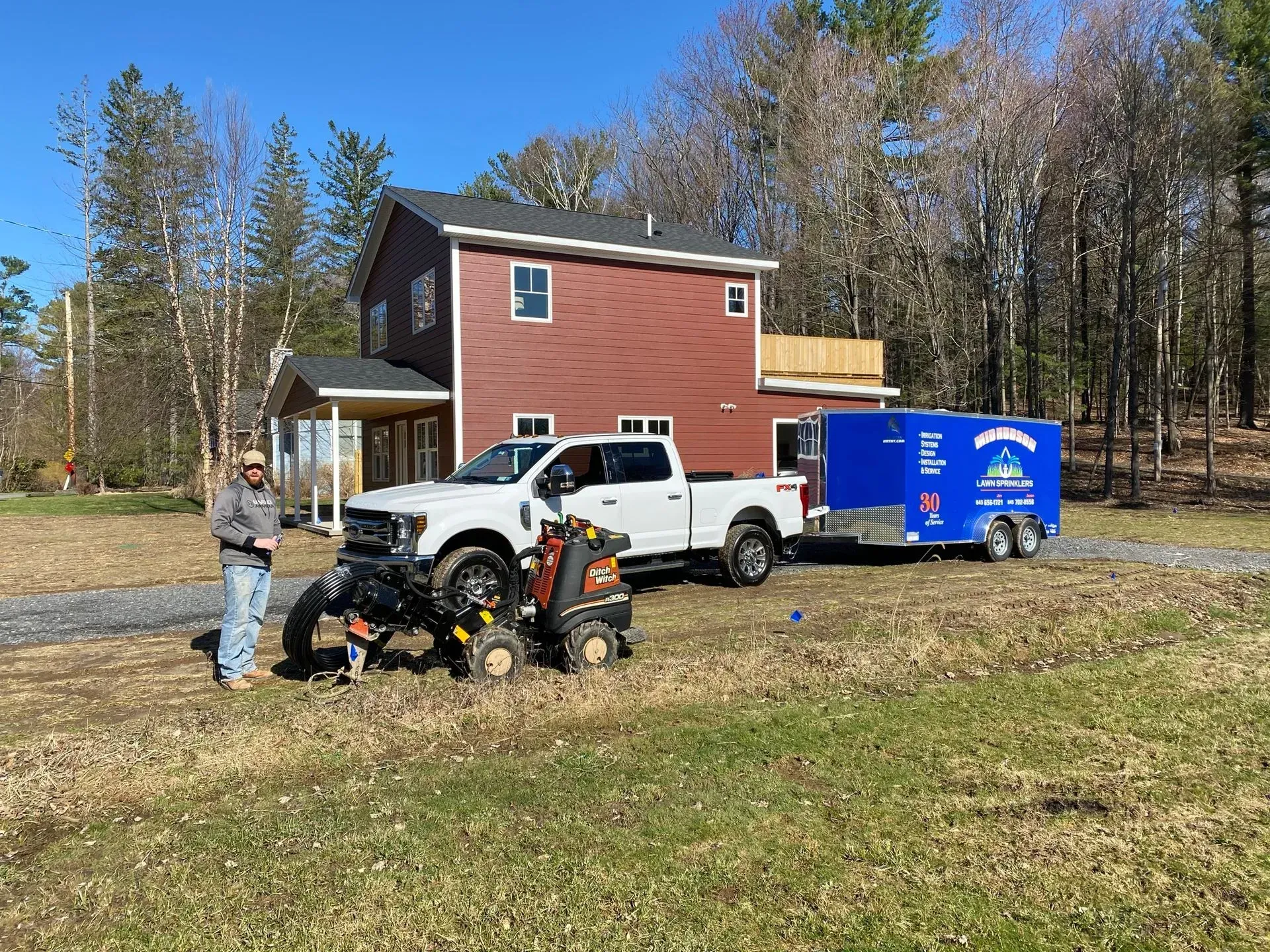 A person stands next to a white pickup truck, an orange equipment skid steer, and a blue trailer in a rural field.
