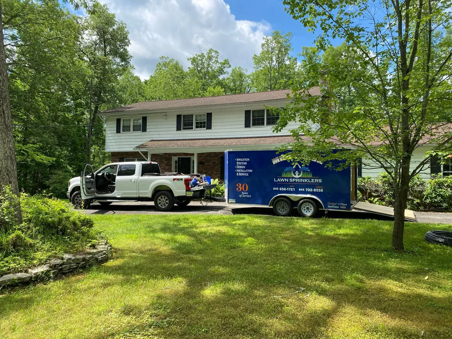 A white pickup truck pulling a blue trailer parked in the driveway of a two-story house surrounded by trees.