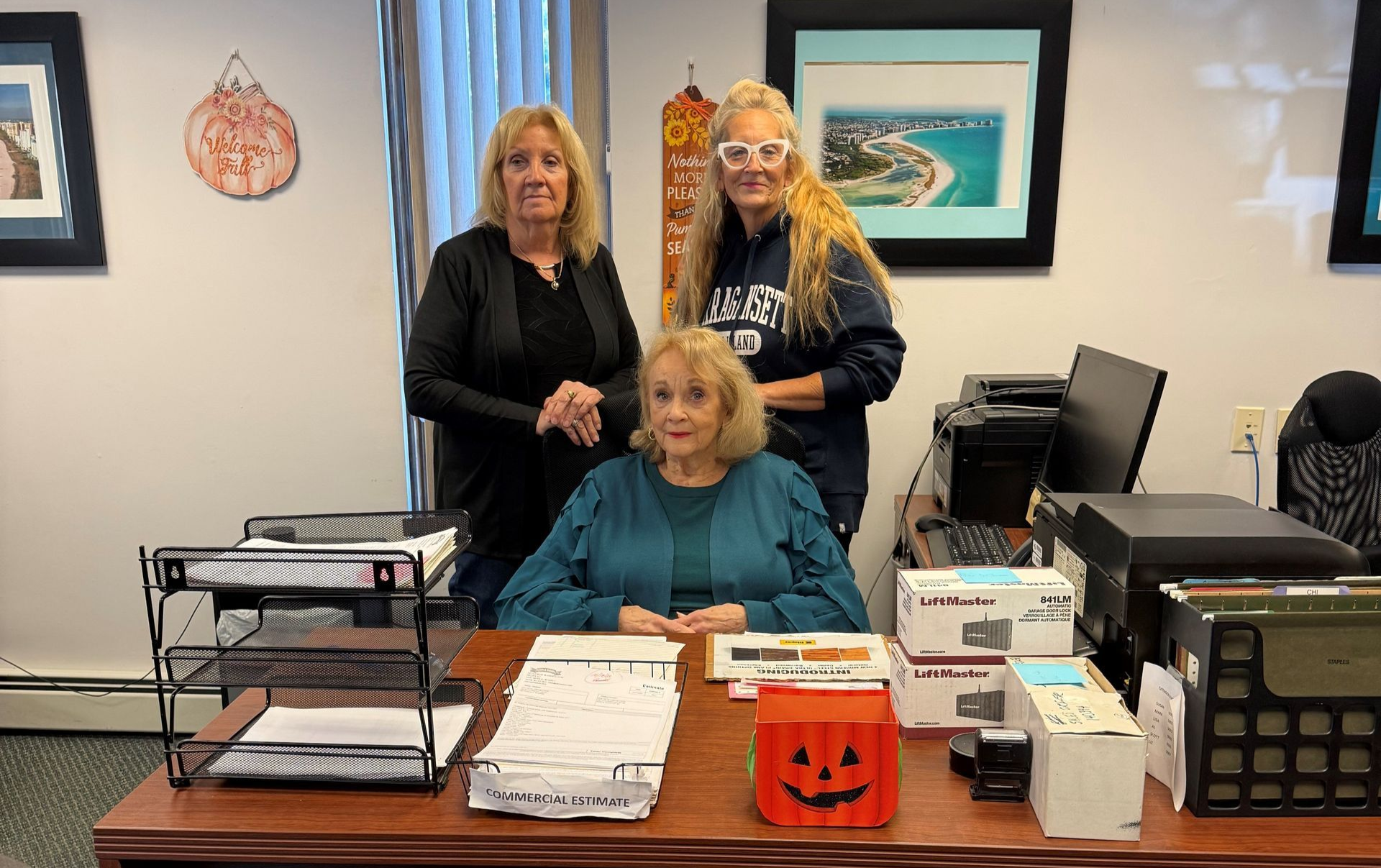 Three women pose in an office, one seated at a desk, two standing behind.