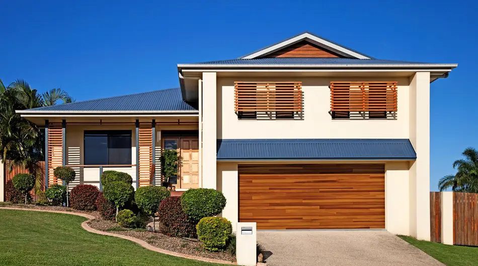 Two-story house with cream siding, wooden accents, blue roof, brown garage door, and a manicured lawn.
