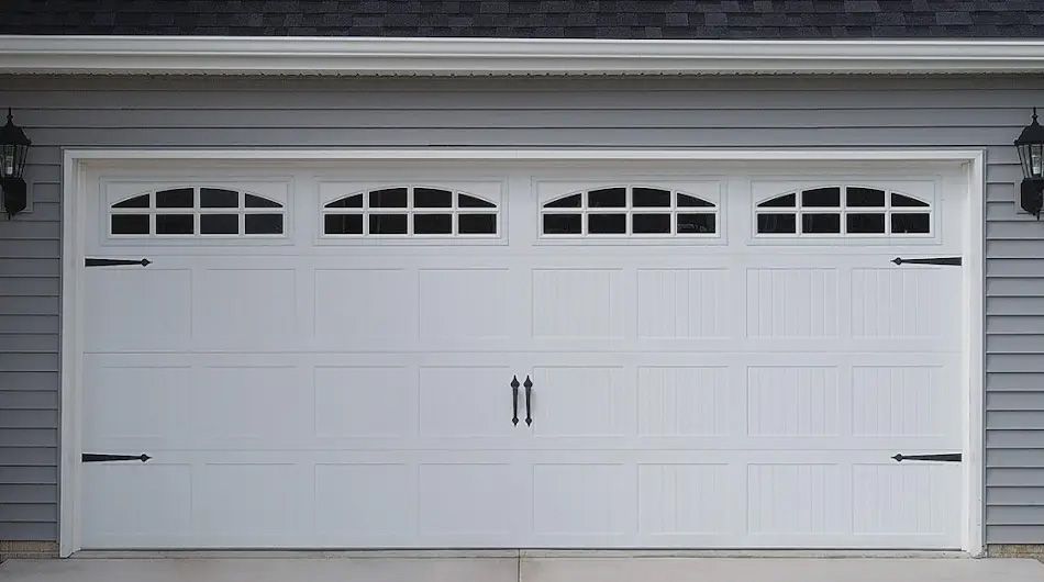 White garage door with arched window panels, black hardware, mounted on a gray house.