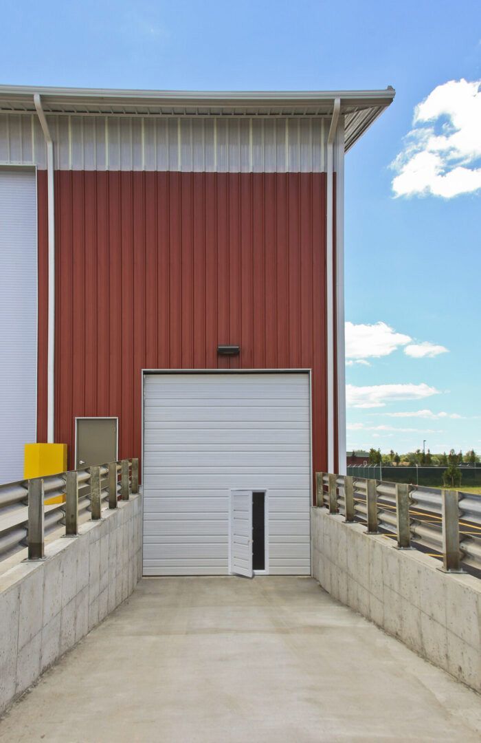 Red and white industrial building with a white loading door and ramp. Blue sky overhead.