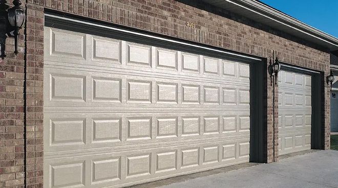 Two tan garage doors on a brick building with sconces and a concrete driveway.