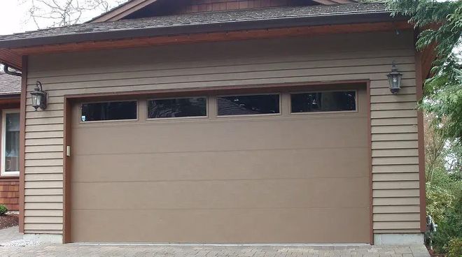 Tan garage door with windows above, flanked by sconces on a building with brown siding.