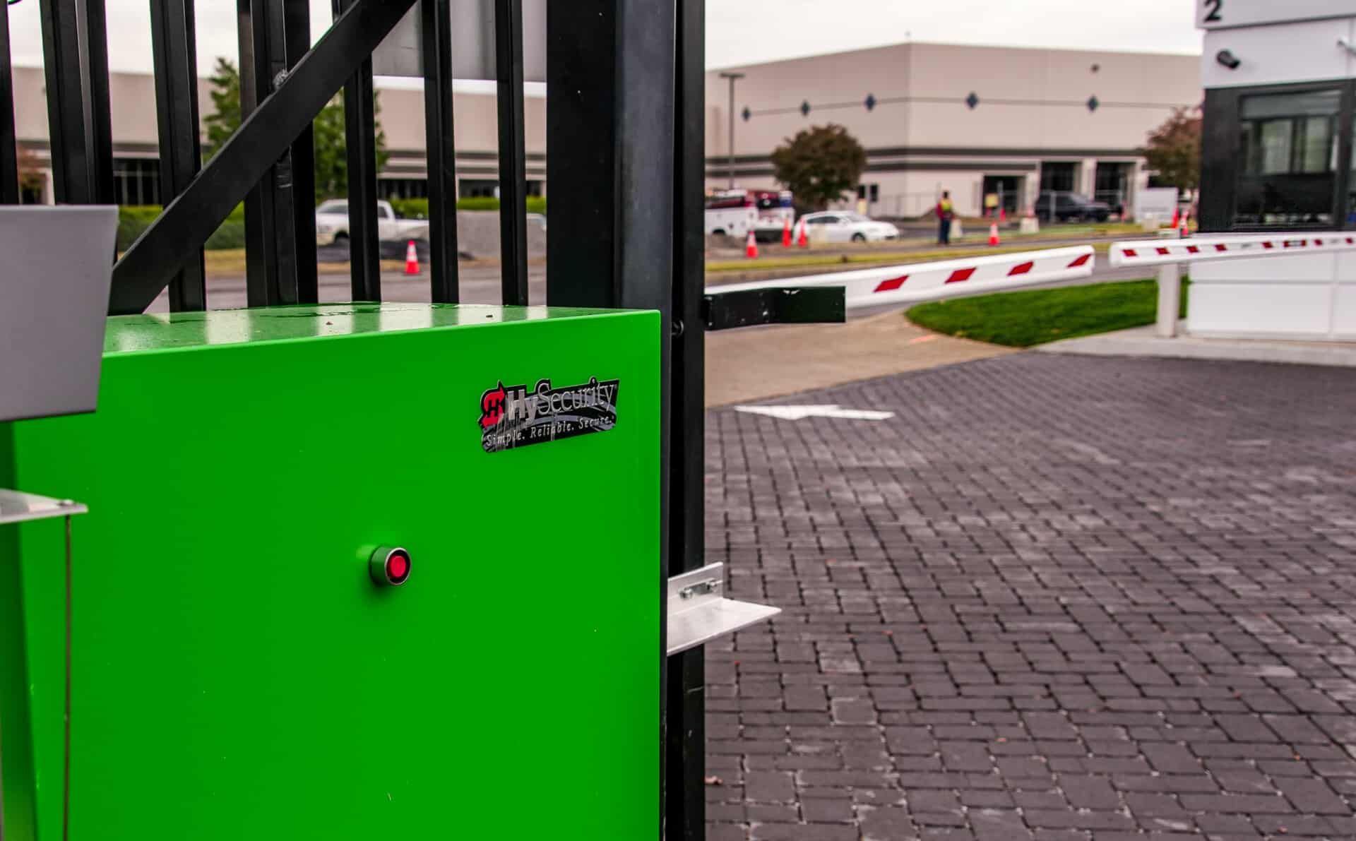 A green box is sitting next to a gate in a parking lot.