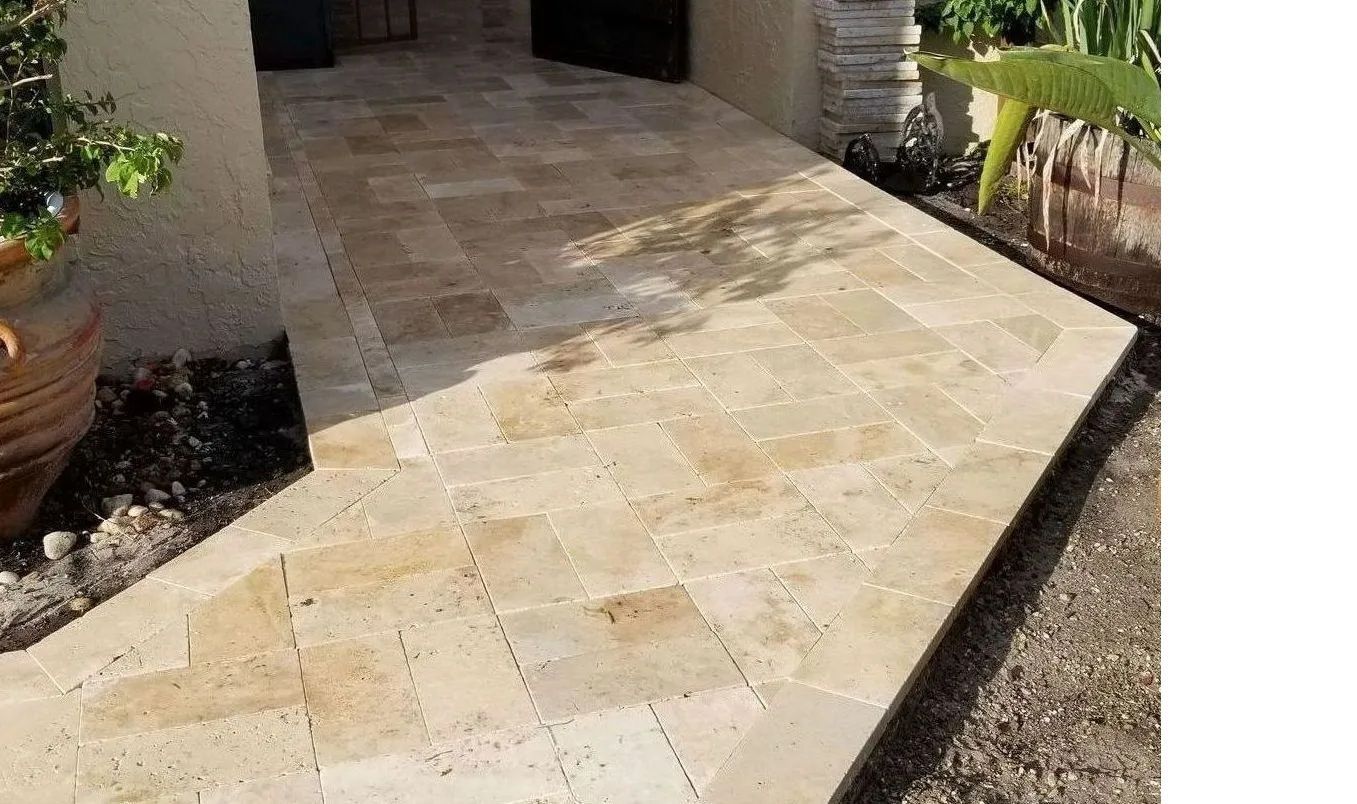 Light tan stone walkway leading to a front porch, bordered by mulch and potted plants.