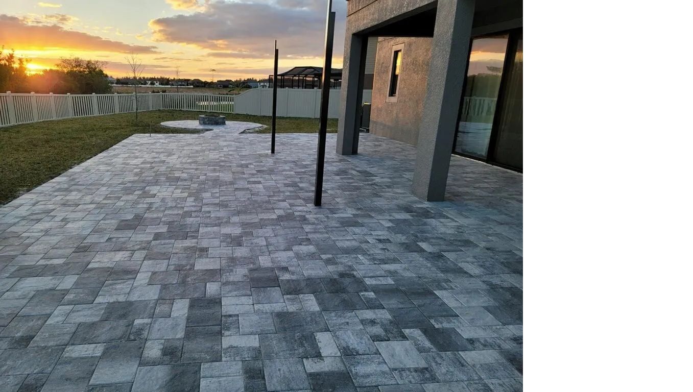 Stone patio beside a modern building at sunset, with a grassy lawn and decorative bridge in the distance