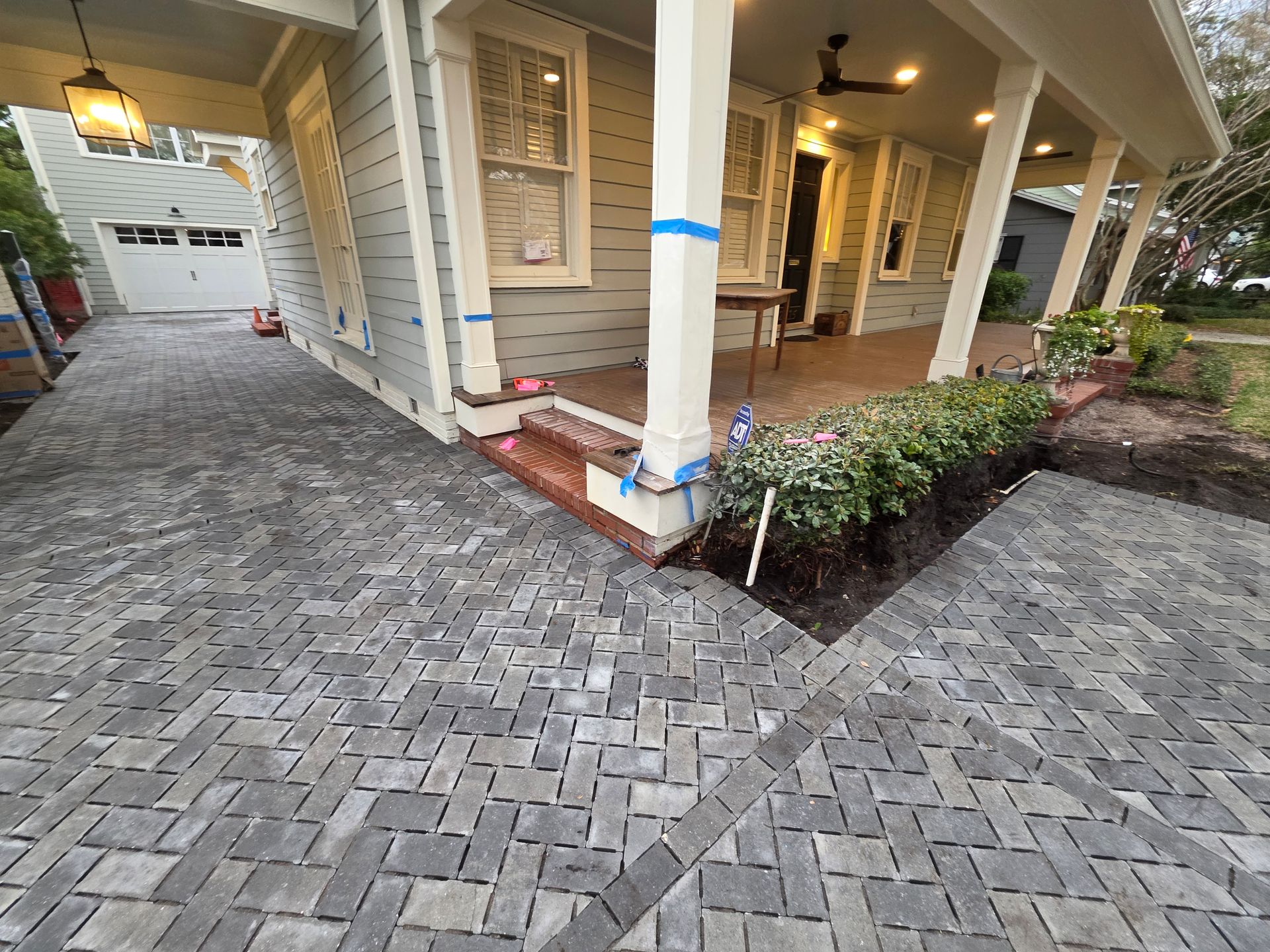 Covered house porch with white columns, blue trim, and a gray brick driveway beside a landscaped garden.
