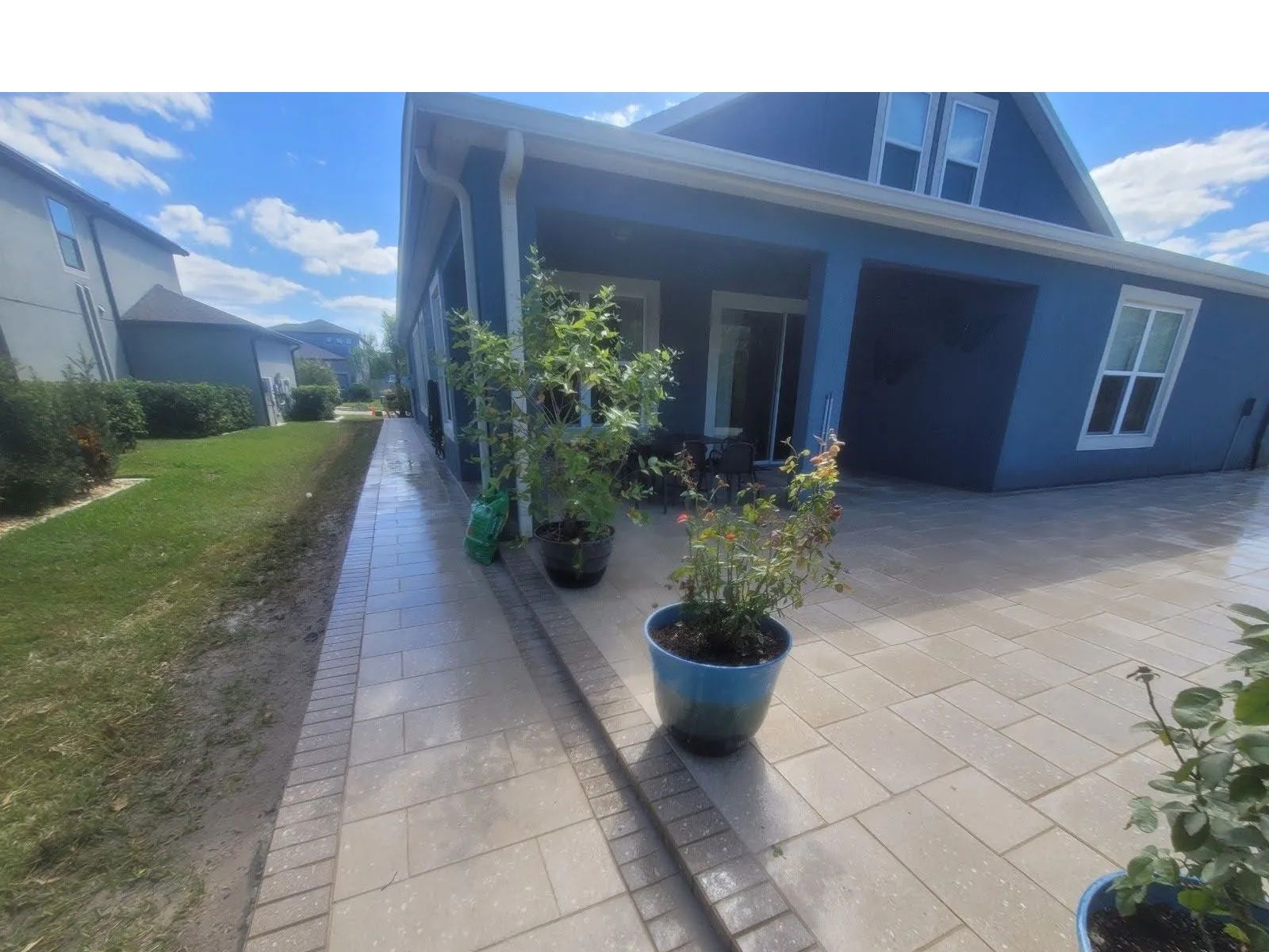 Covered front patio with potted plants beside a blue house and paved walkway