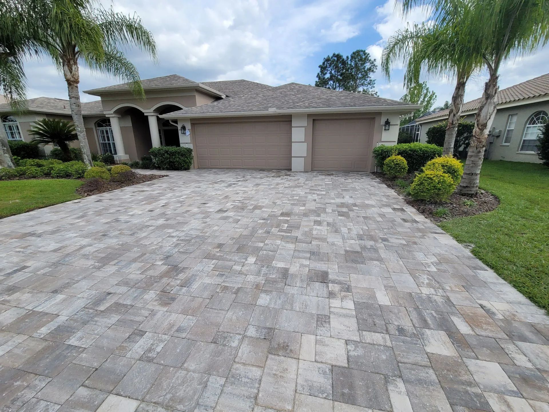 Single-story house with a wide paver driveway, garage, and palm trees in front yard