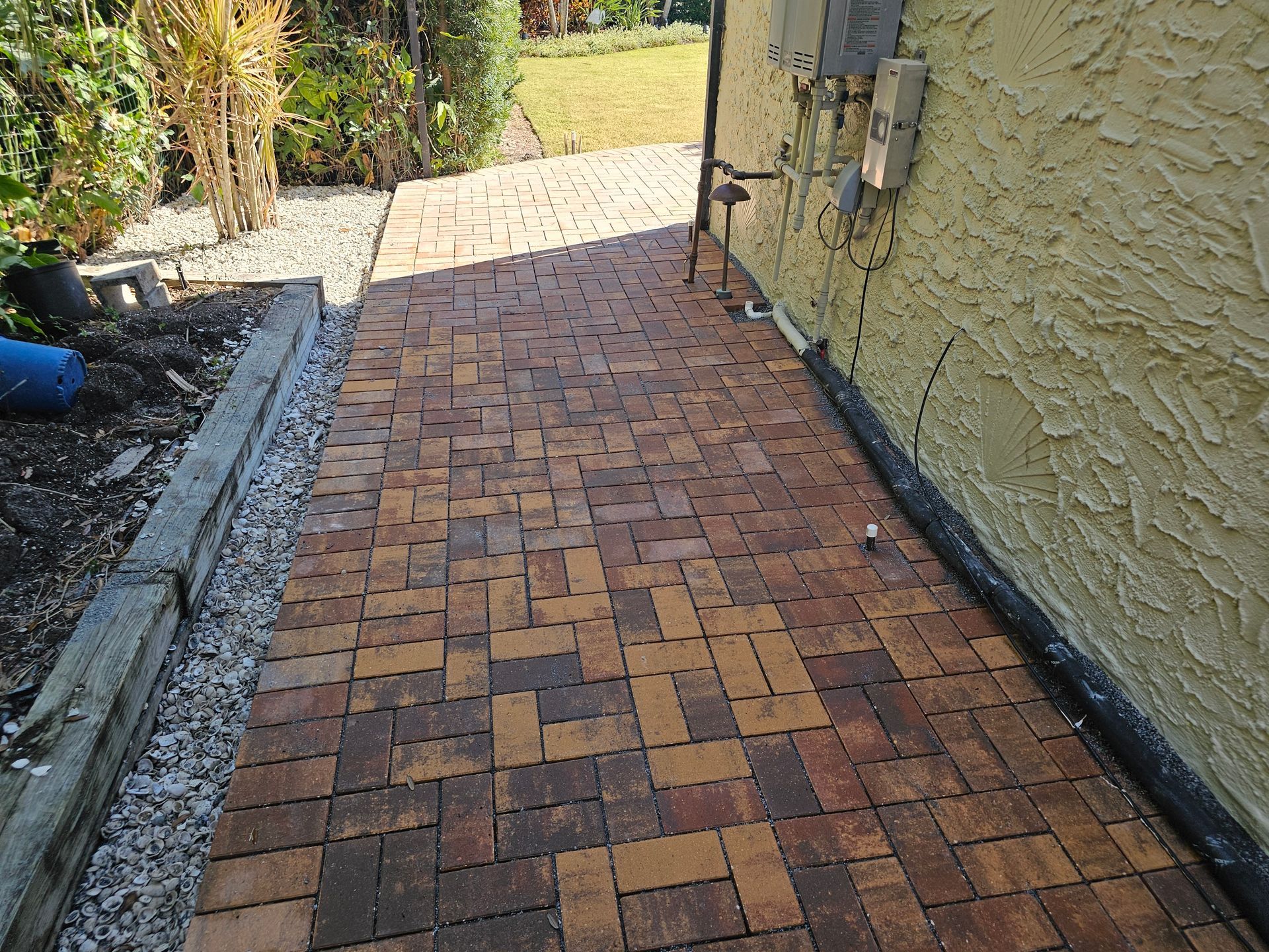 Brick walkway beside a beige stucco house leading to a sunlit yard