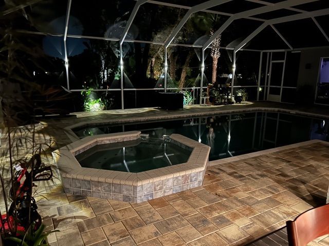 Screened-in backyard pool and hot tub at night with stone patio and tropical landscaping