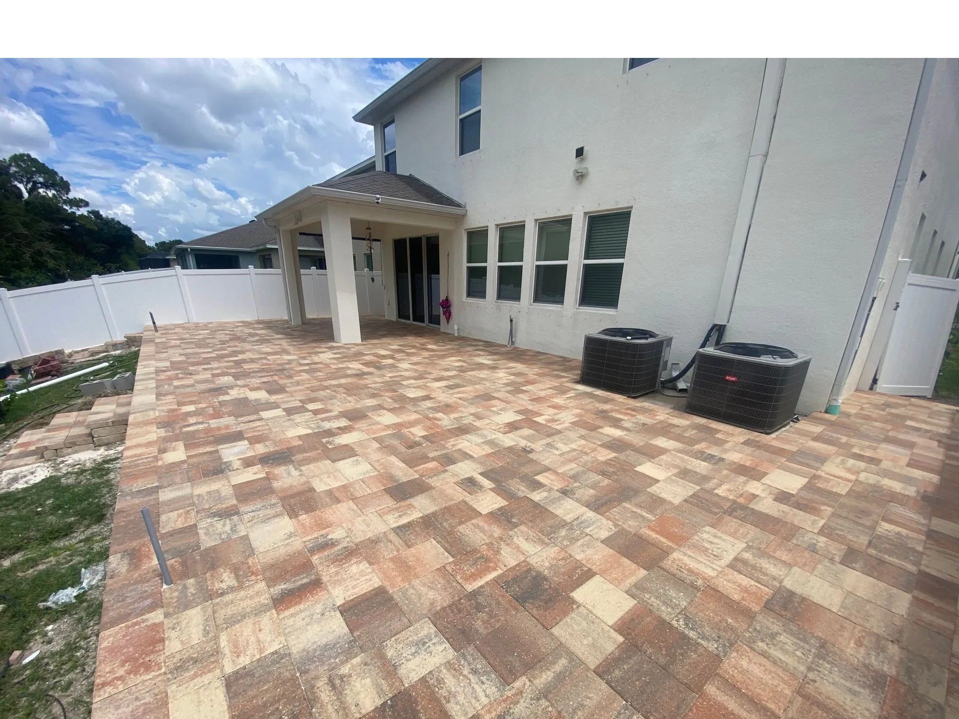 Spacious brick-paver patio beside a white house with windows and a covered entry.