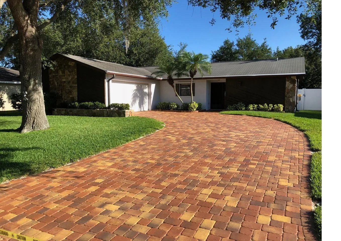 Brick driveway curving to a suburban house with white garage and green lawn under trees