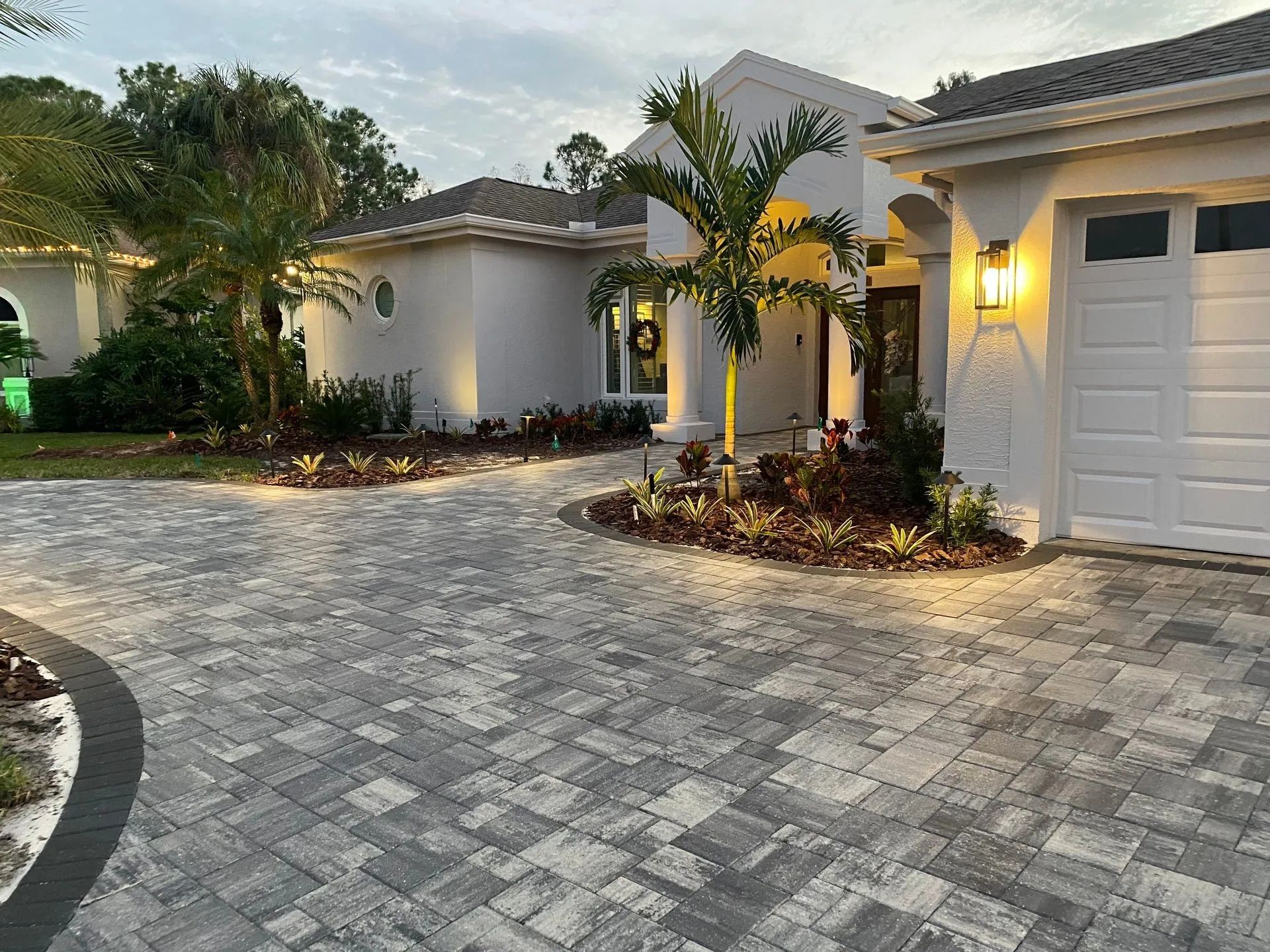 Suburban home driveway with gray pavers, white stucco house, palm trees, and lit exterior lamps at dusk