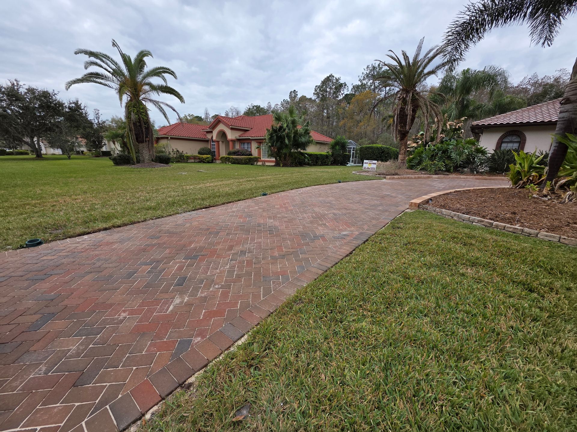 Brick driveway curving through a grassy yard toward a red-roofed house with palm trees under cloudy skies