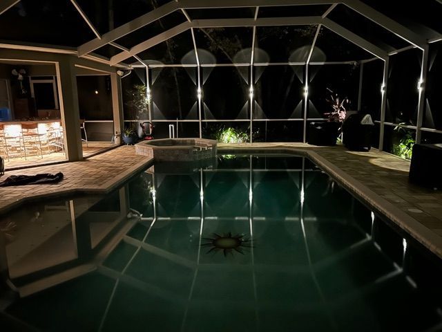 Indoor swimming pool at night with dark water, warm lights, and screened lanai seating area