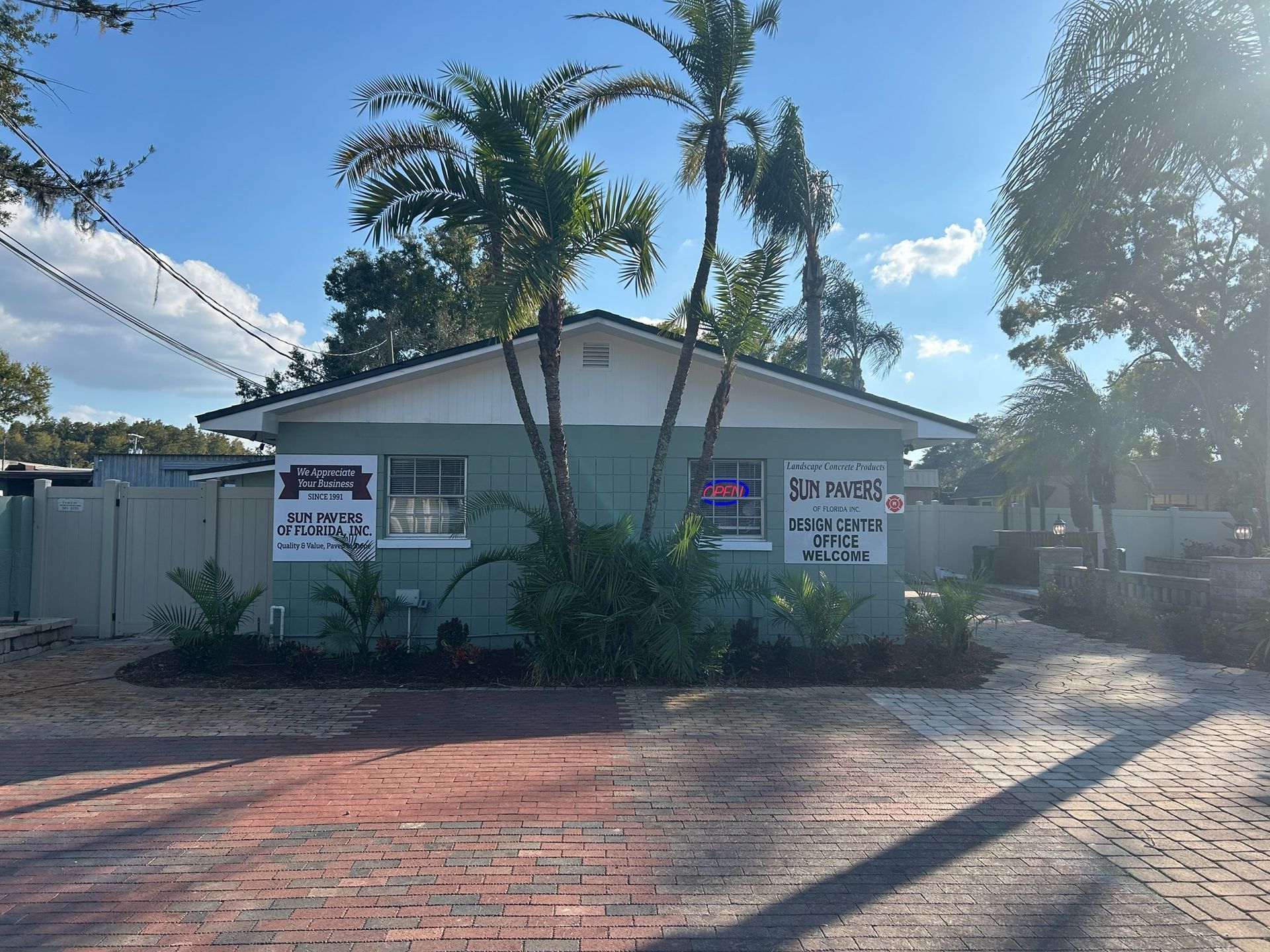 Small white house with palm trees, signs on the front, and a sunlit driveway under a blue sky.