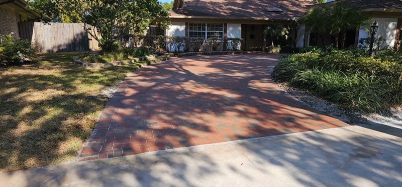 Brick driveway leading to a house with trees and shrubs in front-yard shade