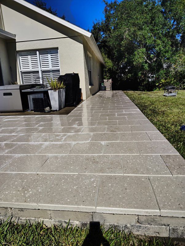 Sunlit tiled patio beside a house, with barbecue grills and a grassy yard on the right.