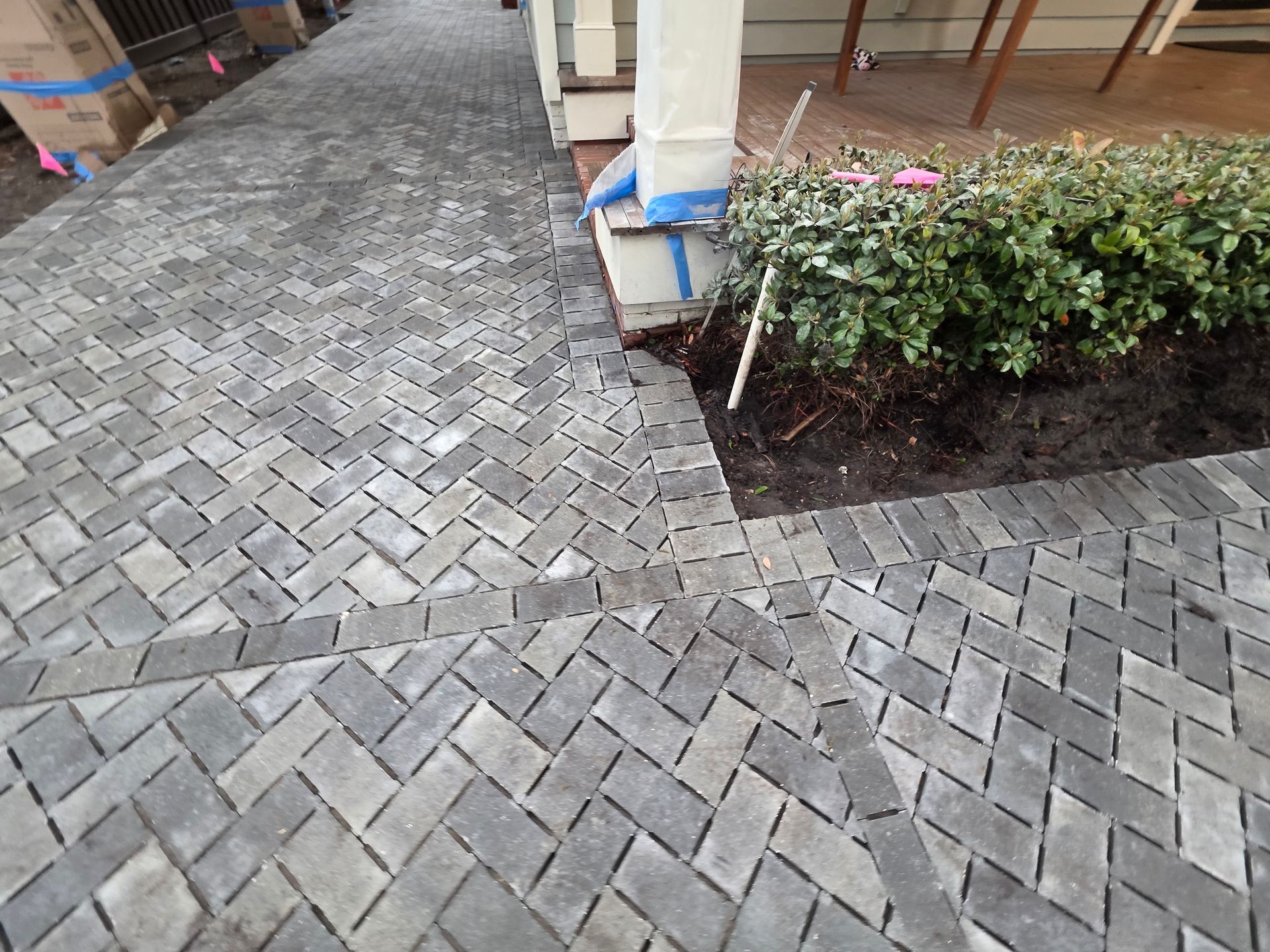 Gray brick-paved walkway beside a building, with a small garden bed and trimmed shrubs on the right.