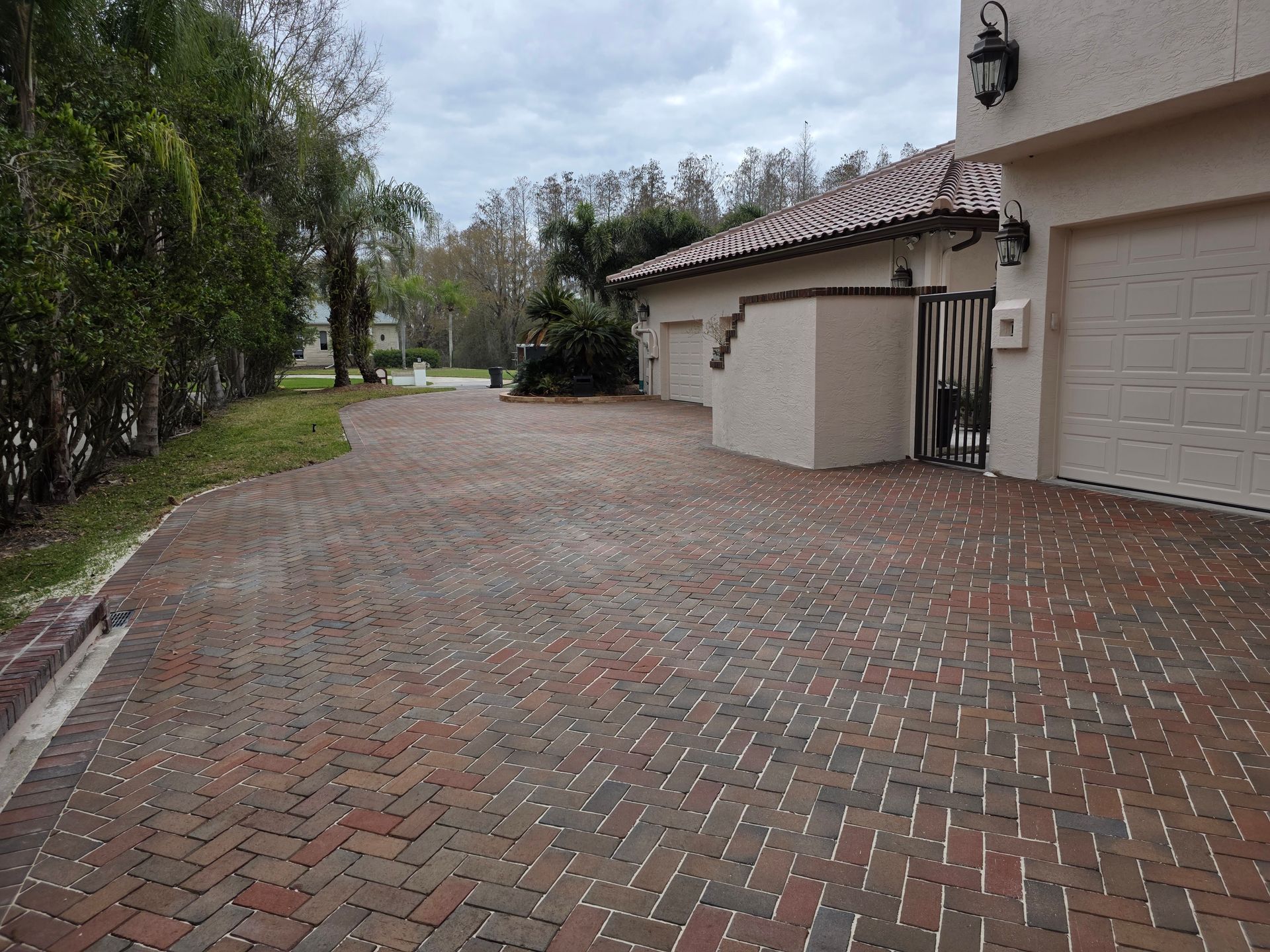 Brick driveway beside a house and garage, bordered by trees and shrubs on a cloudy day.