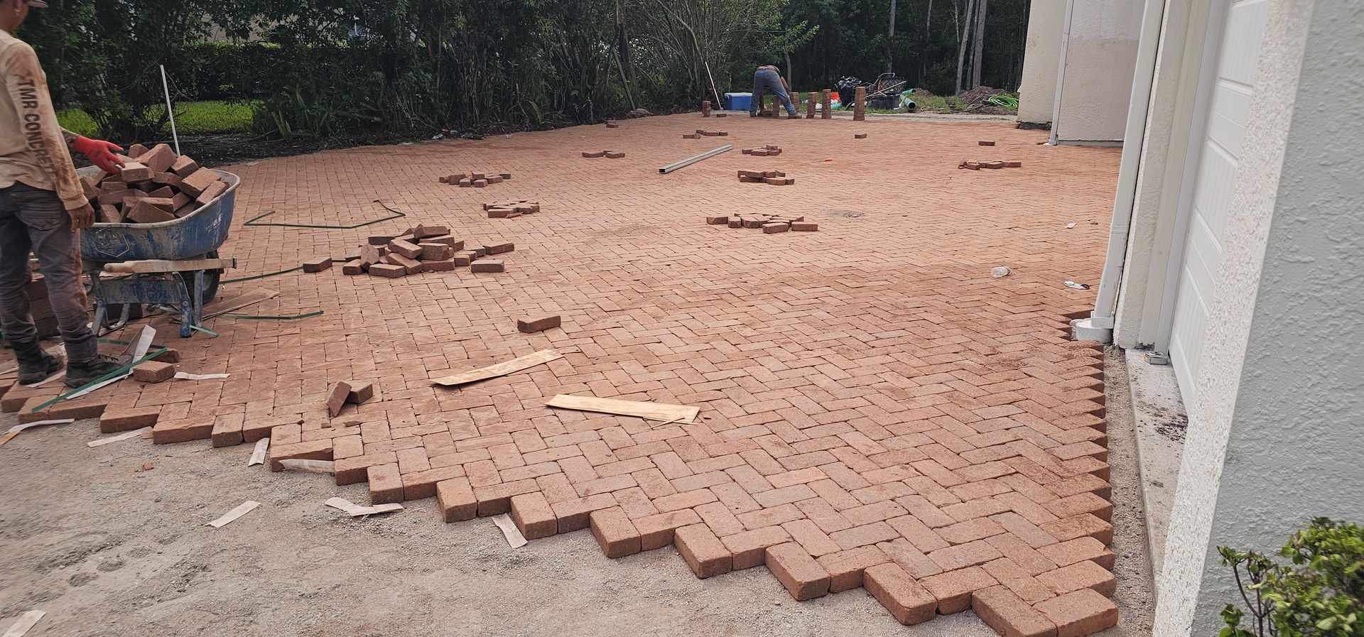 Partially installed red-brown patio pavers beside a house and wall, with scattered materials and debris
