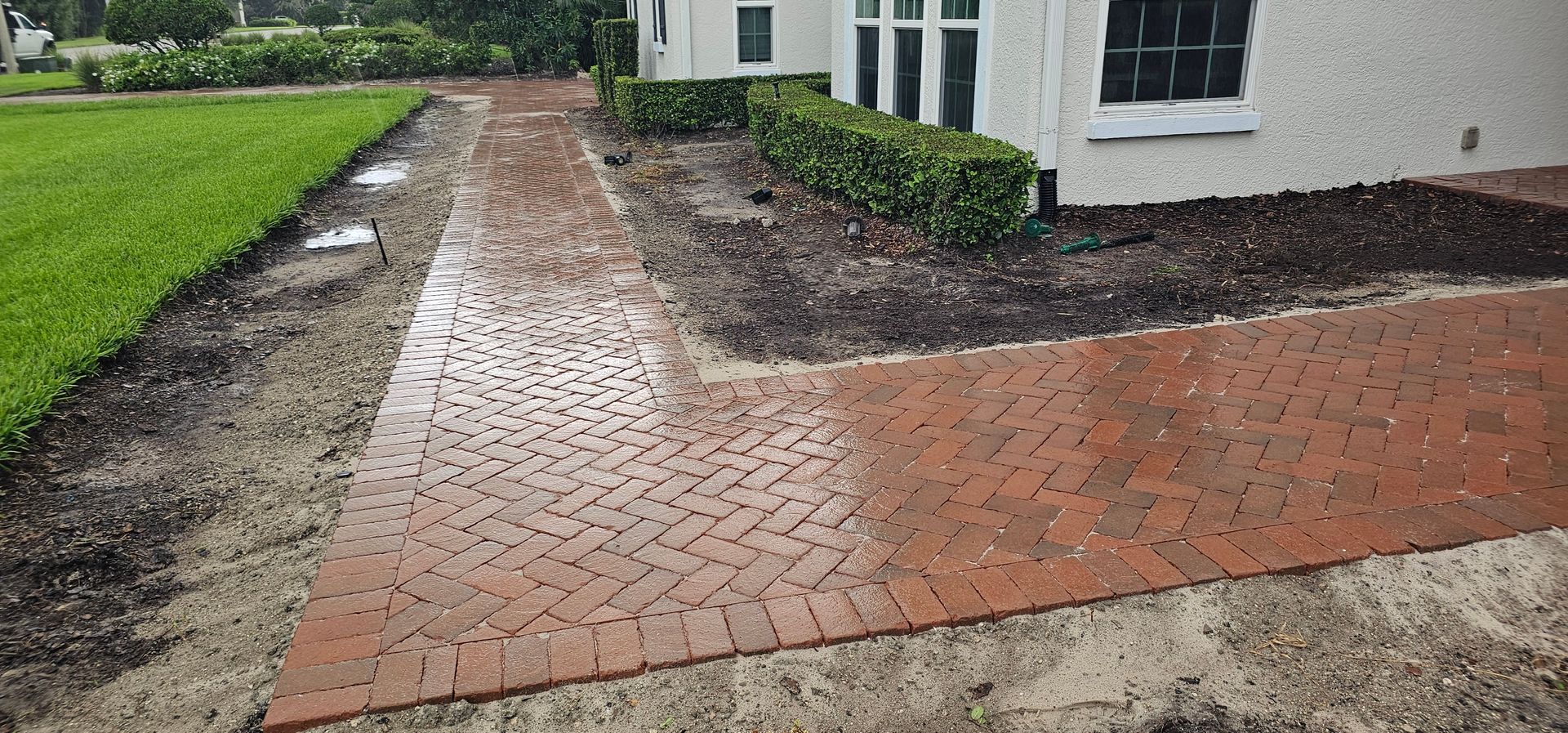 Brick walkway beside a white house, bordered by grass and bare soil, leading into a driveway area.