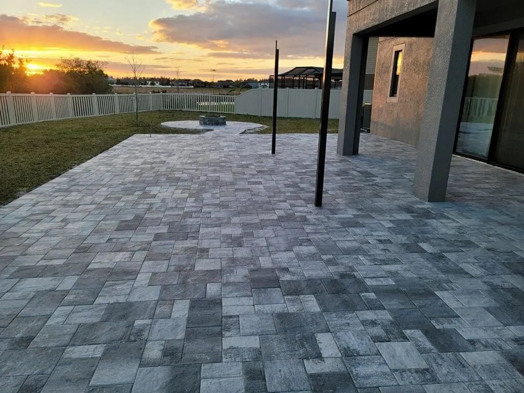 Modern stone patio at sunset beside a house, with a lawn, railing, and ocean view.