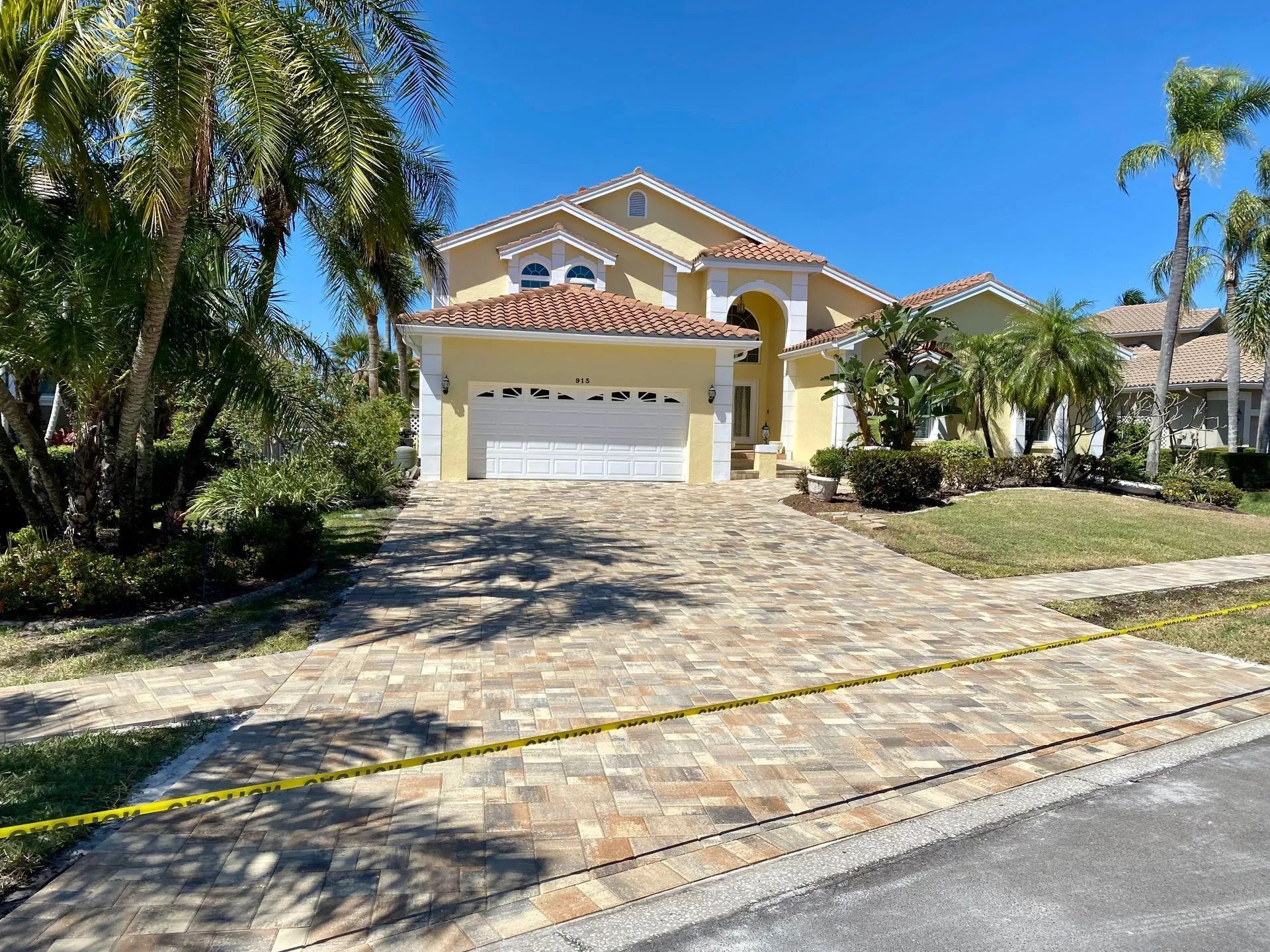 Large yellow stucco house with a white garage, tiled driveway, and palm trees under a blue sky