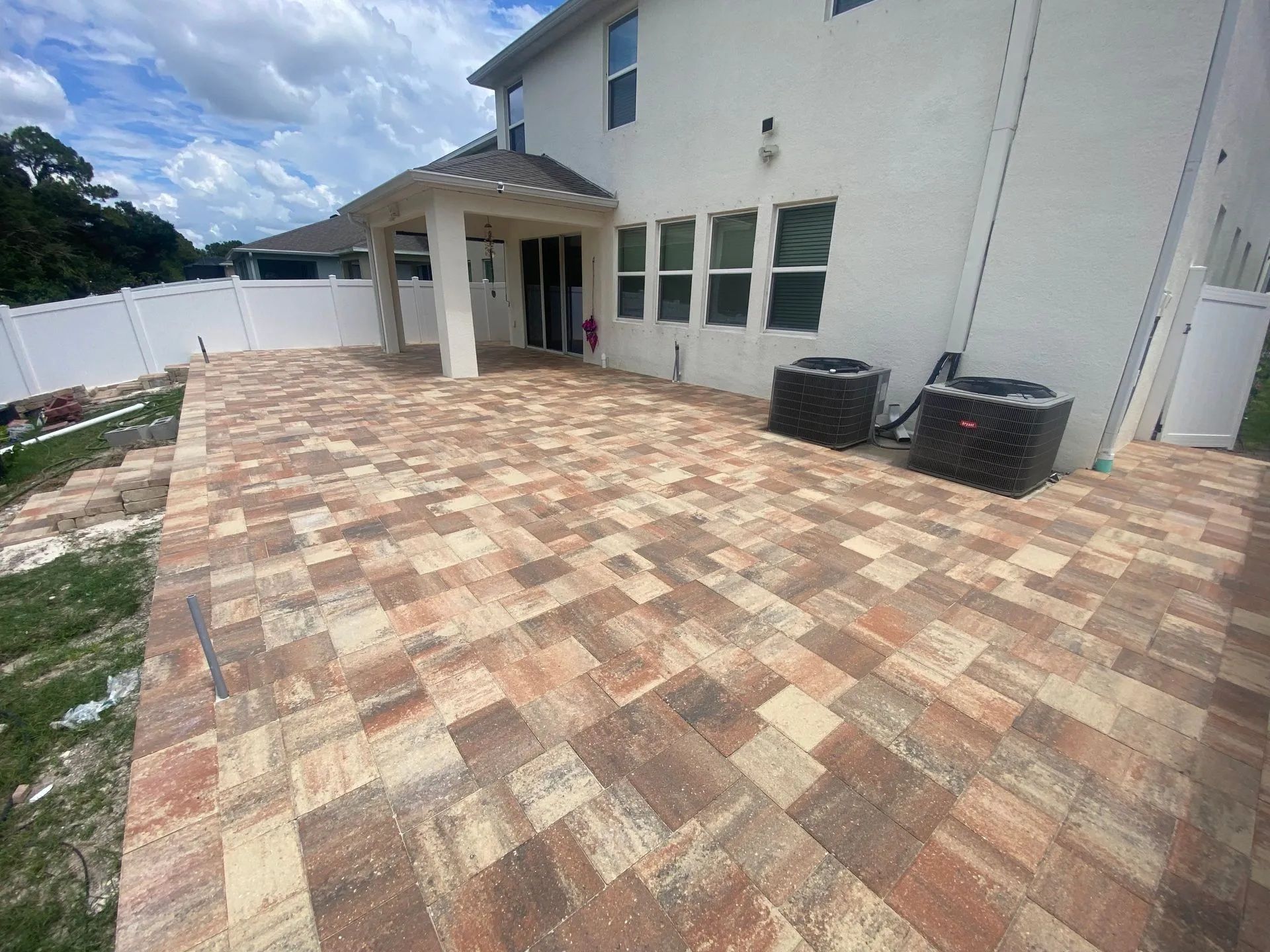 Backyard patio with tan brick pavers beside a white house and covered entryway