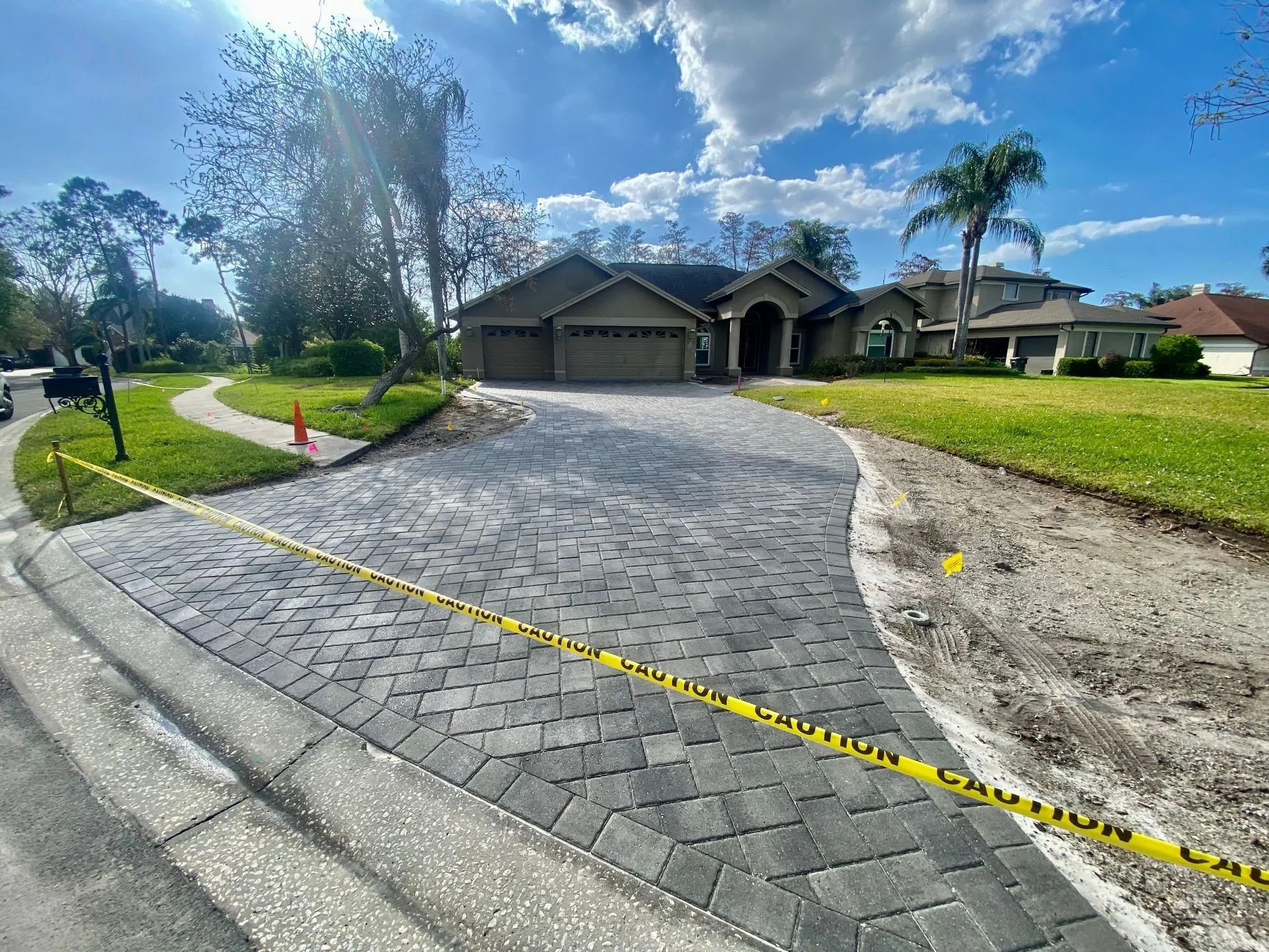 Suburban driveway with paver border, yellow caution tape, and houses under a bright blue sky