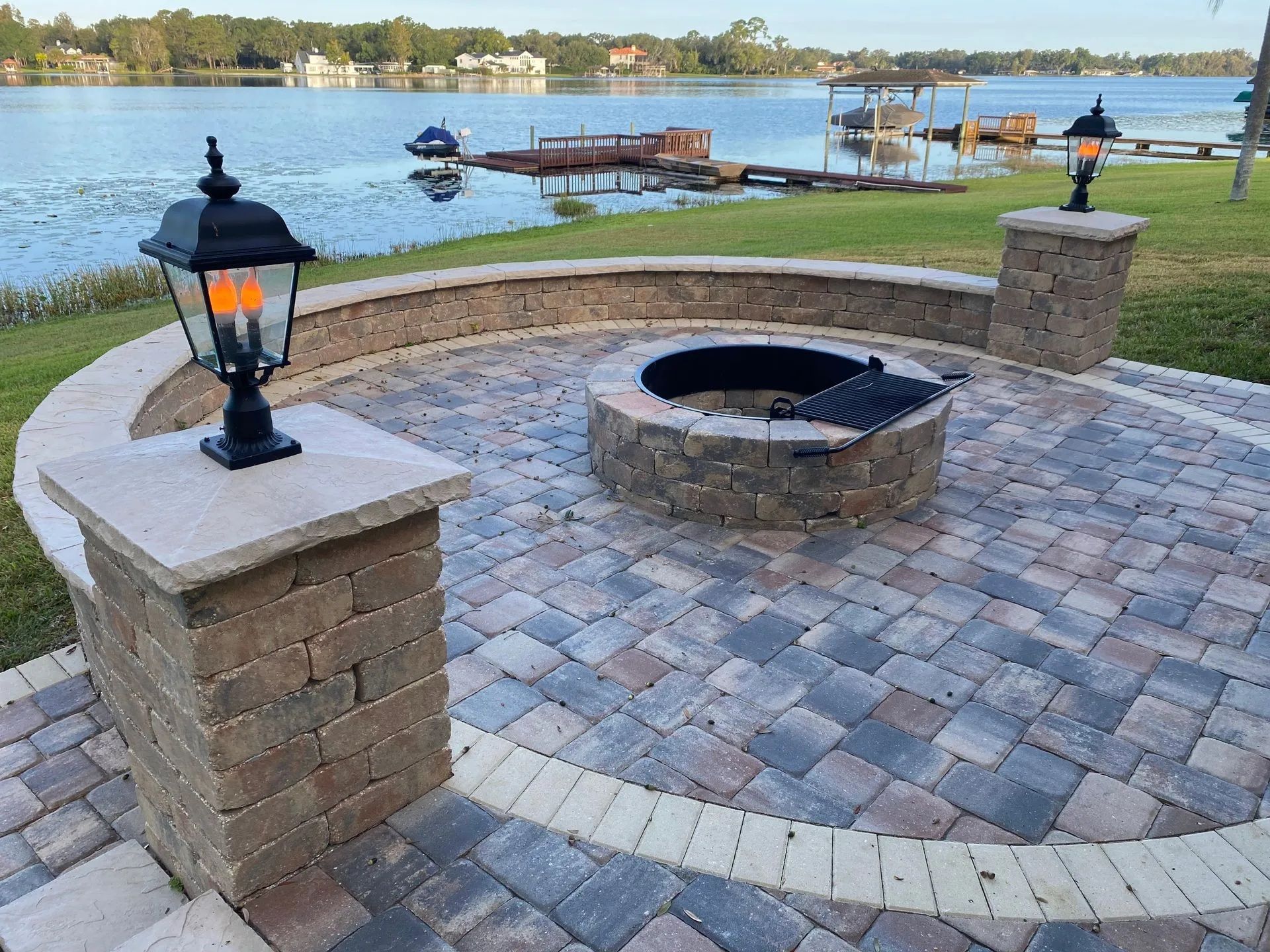 Brick fire pit beside a lakeside patio with black lanterns and a grassy shoreline