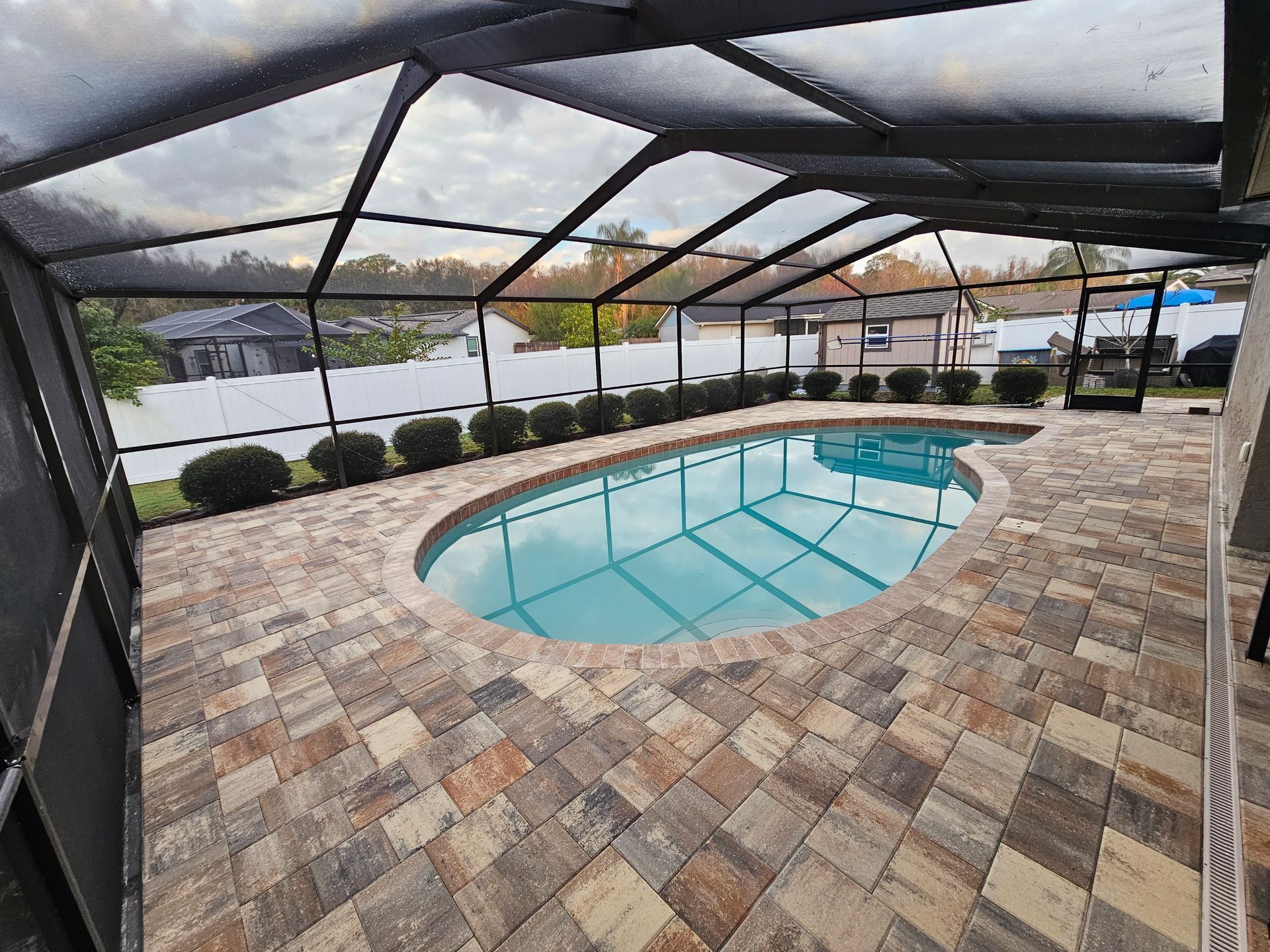 Screened-in patio with a small oval pool and stone pavers, overlooking a fenced yard and road.