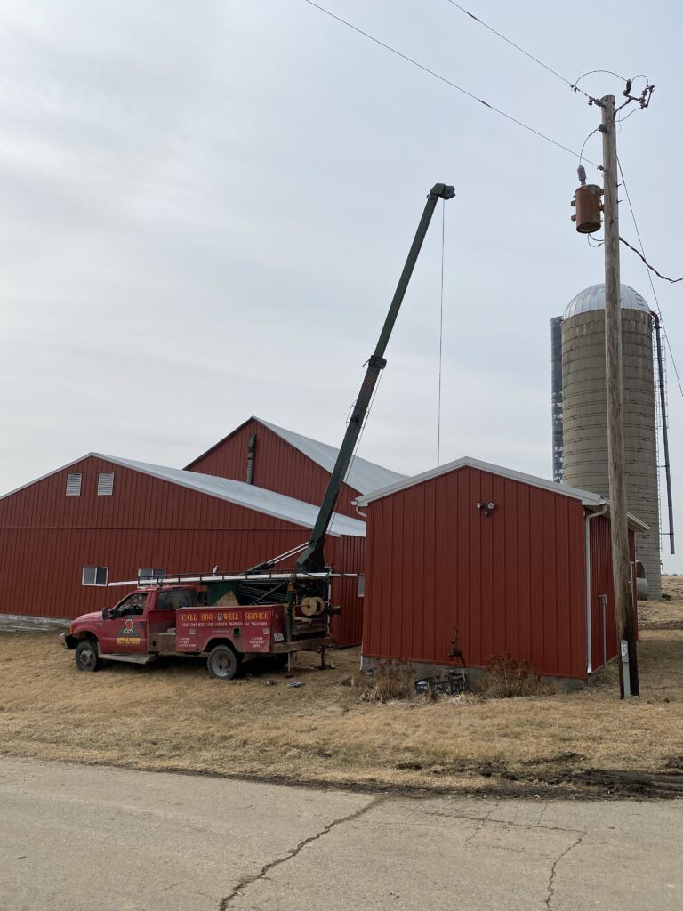 Red crane truck beside red farm buildings, with a tall grain silo in the background.
