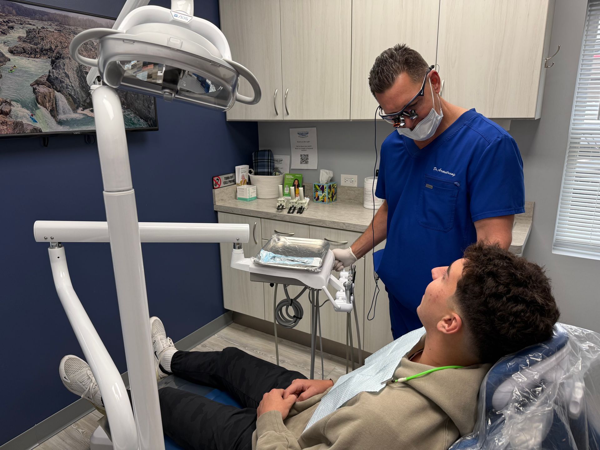 Dentist examining a patient in a dental office; dentist wears mask and scrubs, patient reclined.