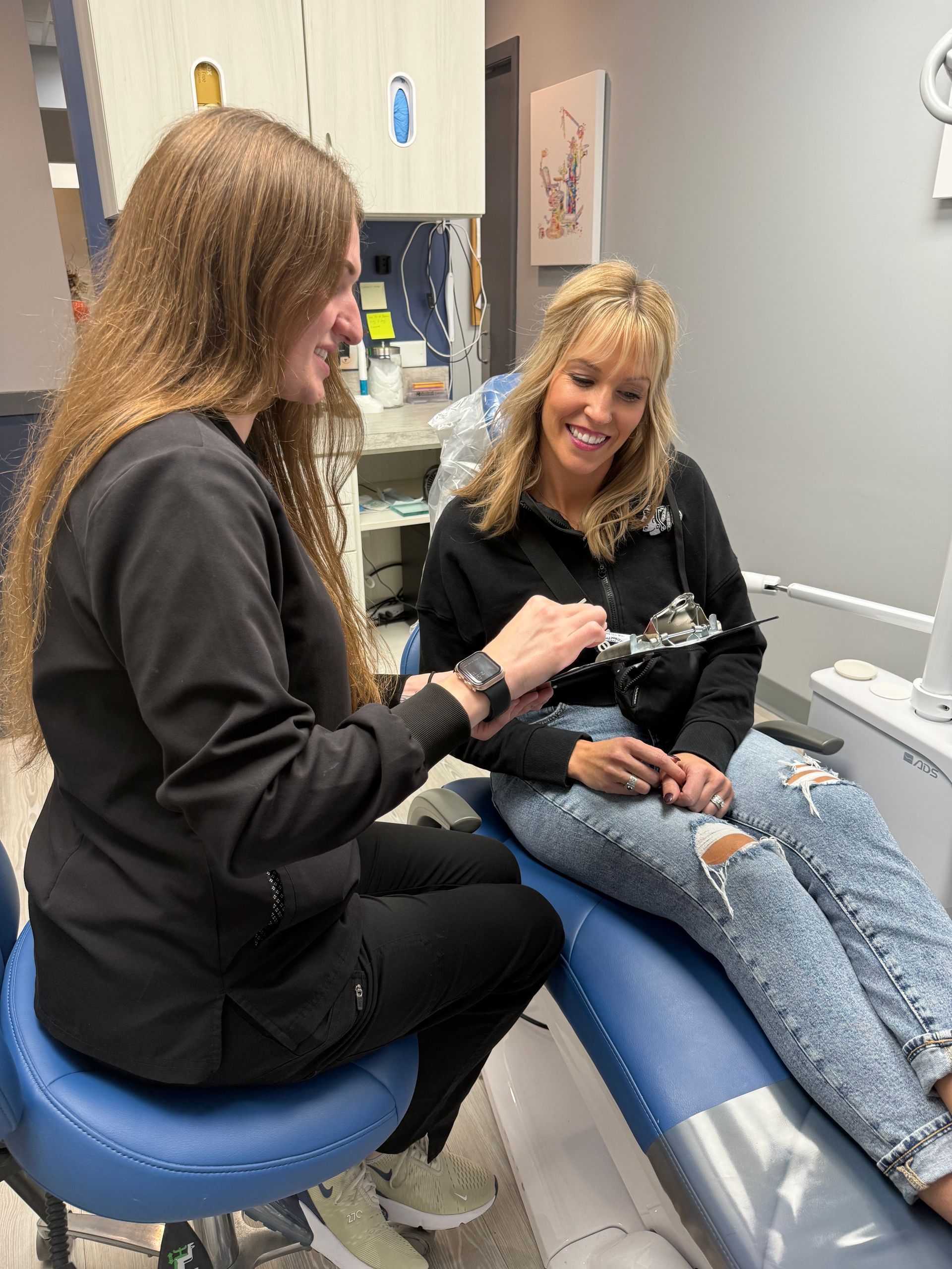 Dental assistant showing tool to seated patient in a dental office. Both are smiling.