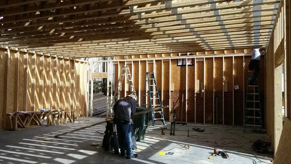 Construction site: interior view of a room with wooden frames. Two workers on ladders, one in front of a green bin, and open doorway.