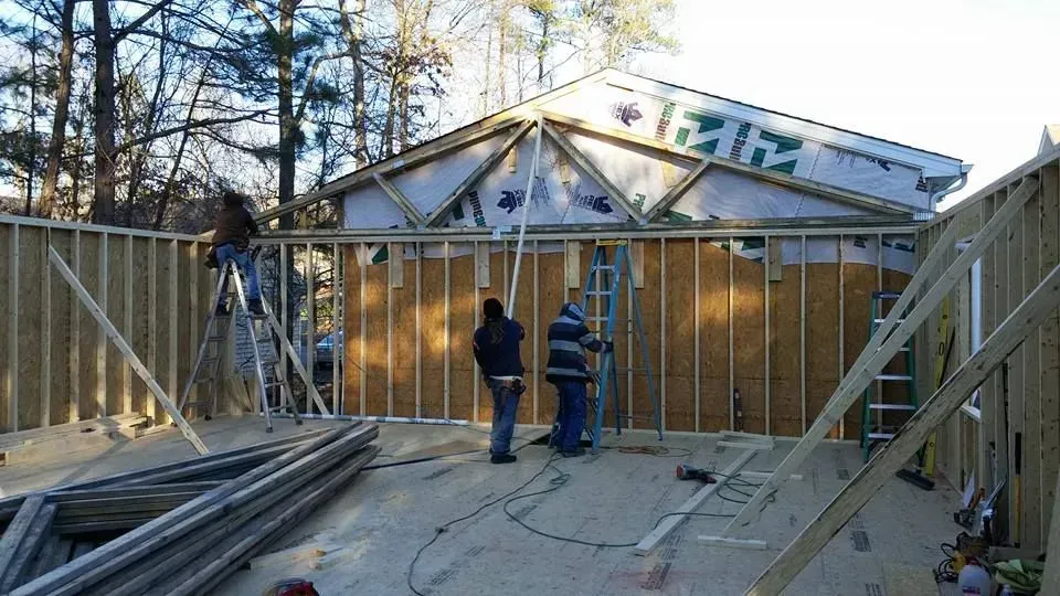 Construction workers framing a house's walls, roof rafters visible. Outdoors, with OSB sheathing and blue tarp.