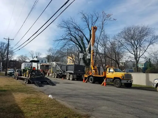 Crews working on power lines. Trucks and equipment line the street near bare trees and homes.