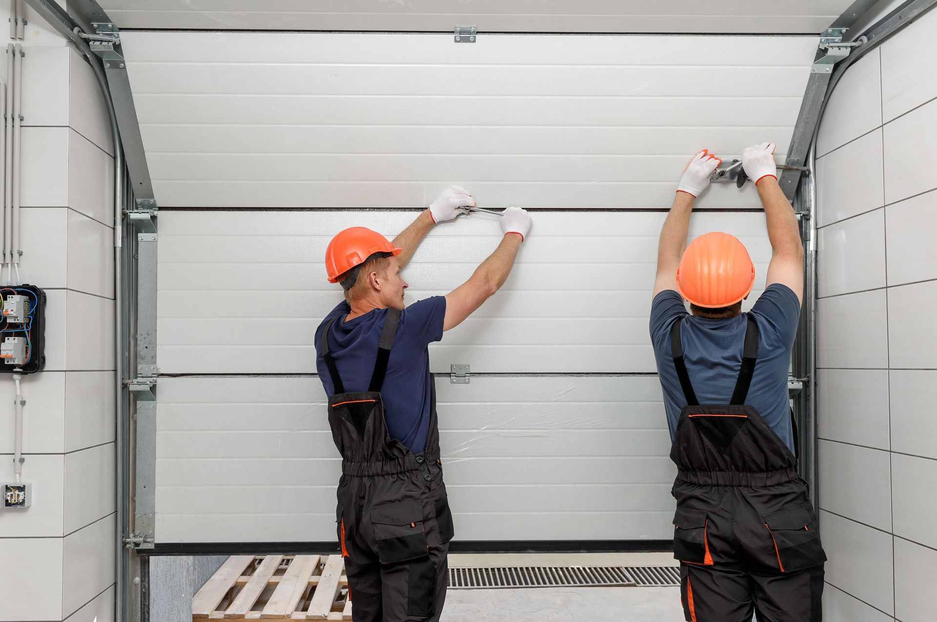 Two workers in hard hats installing a white garage door in a warehouse.