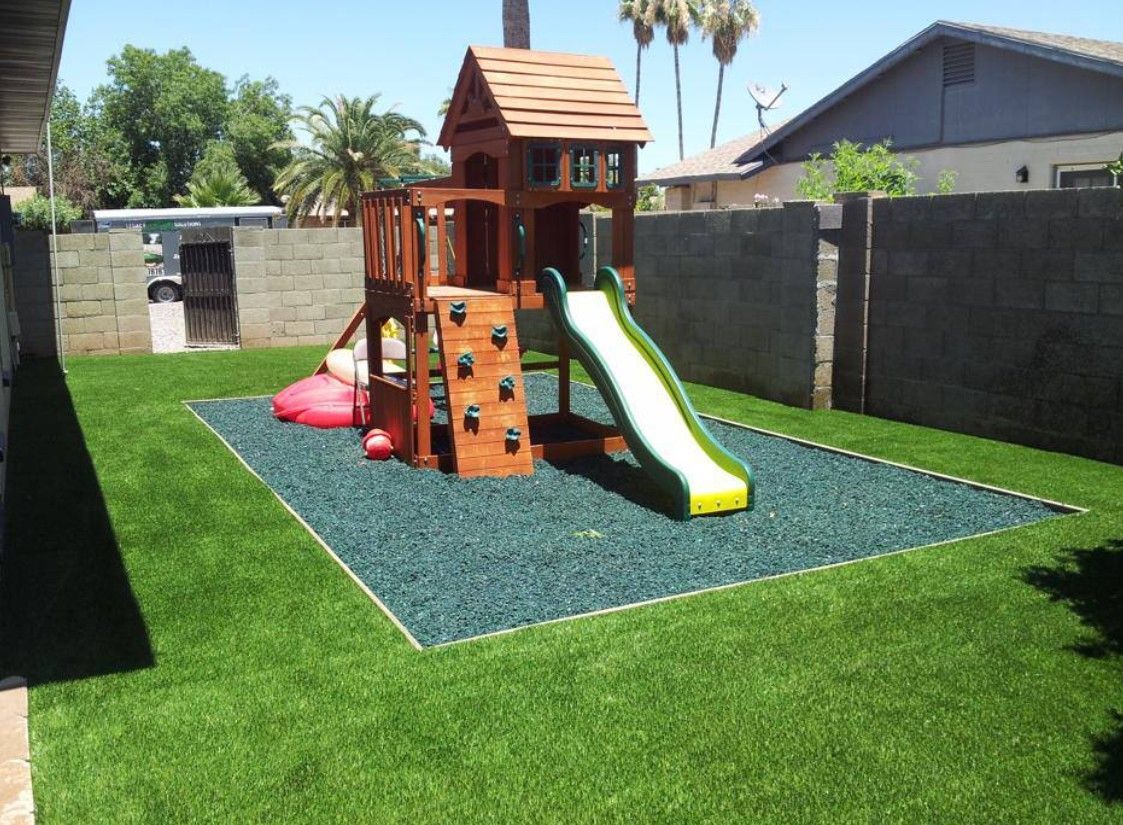 Backyard playground with a wooden playset, slide, and climbing wall on green grass and blue mulch.